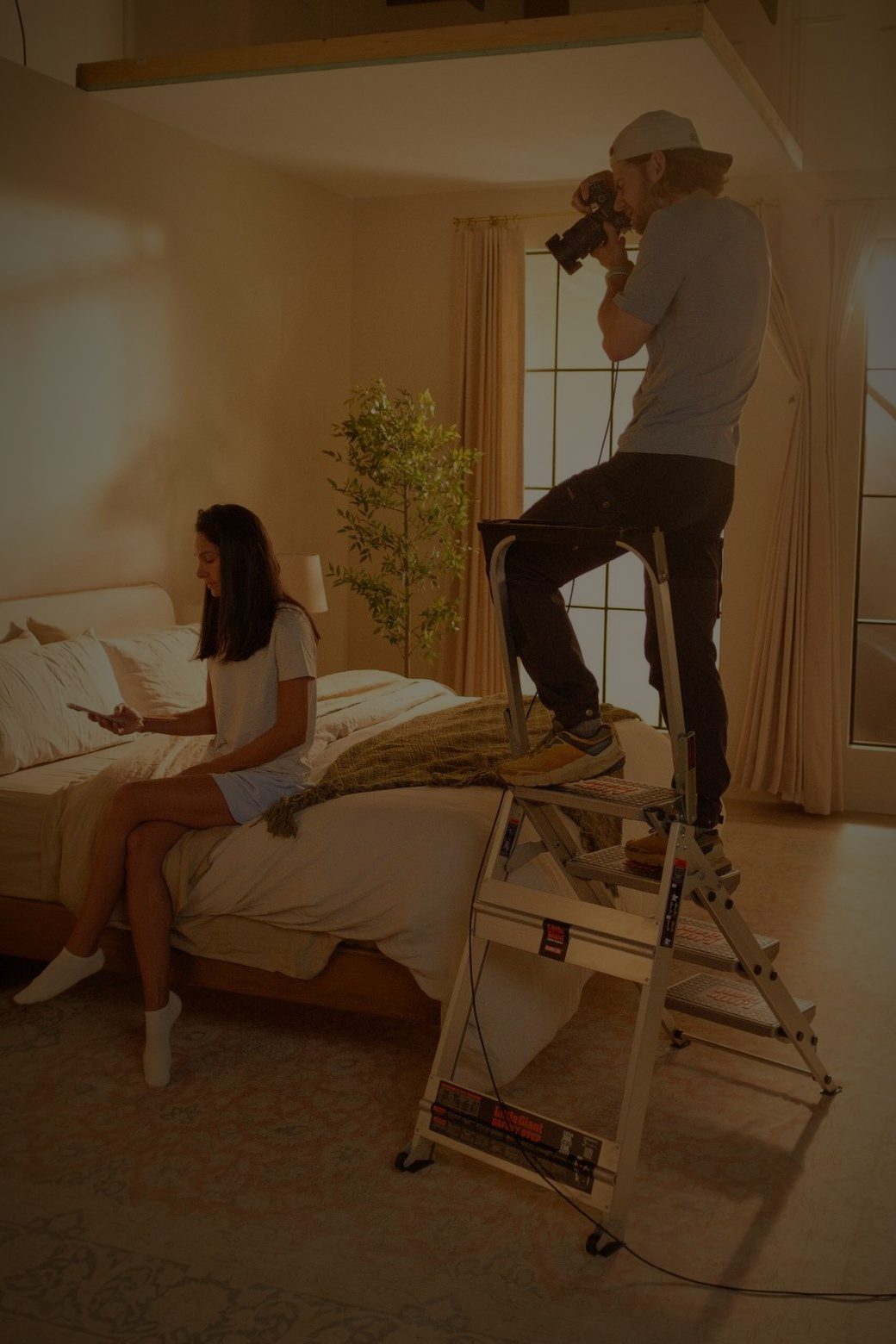 A photographer on a step ladder shooting a woman seated on a bed on a dressed studio set.