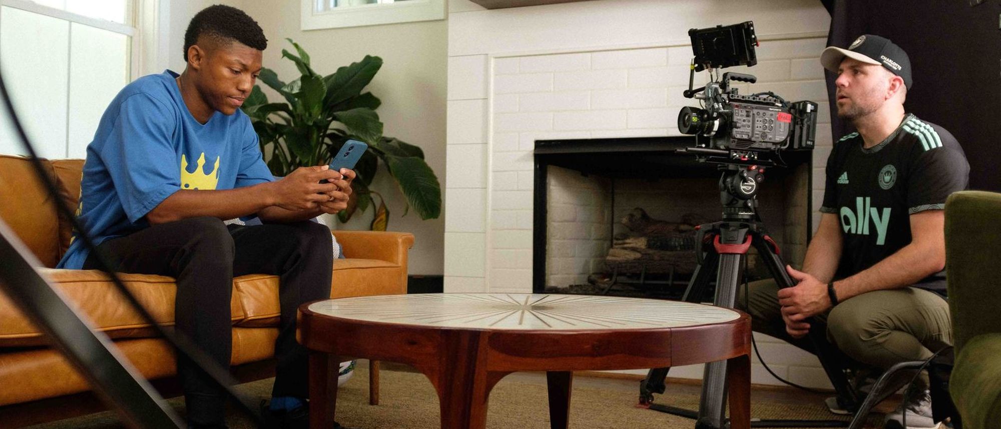Two people seated in a living room set during an interview setup, with a camera on sticks between them.